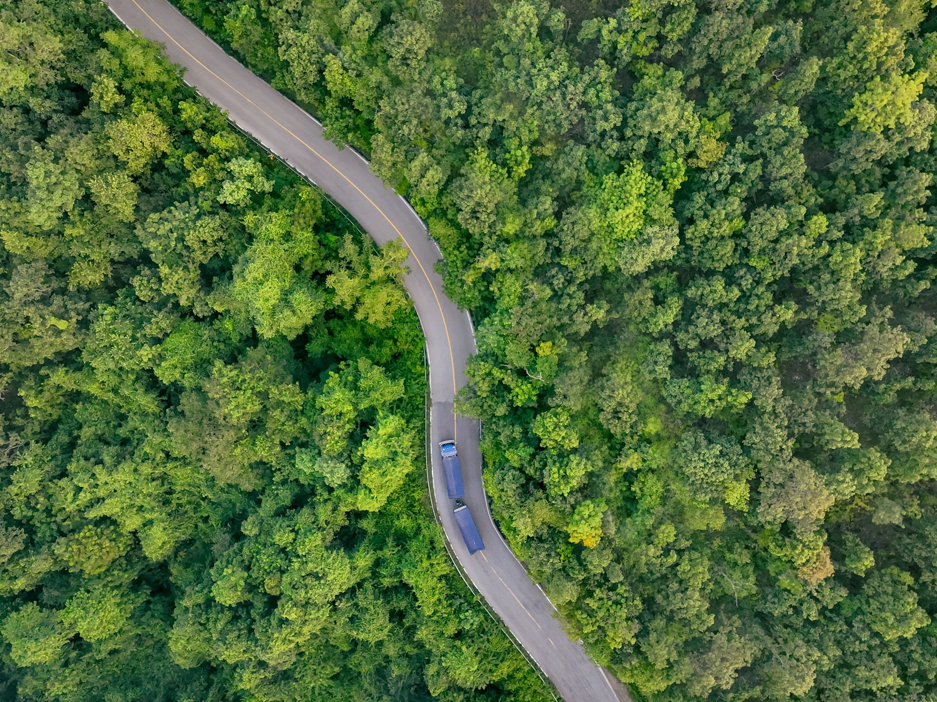 Aerial view hydrogen truck on forest highway. Sustainable transportation. Blue zero emission vehicle driving through green landscape. Eco-friendly logistics solution. Clean energy freight delivery.